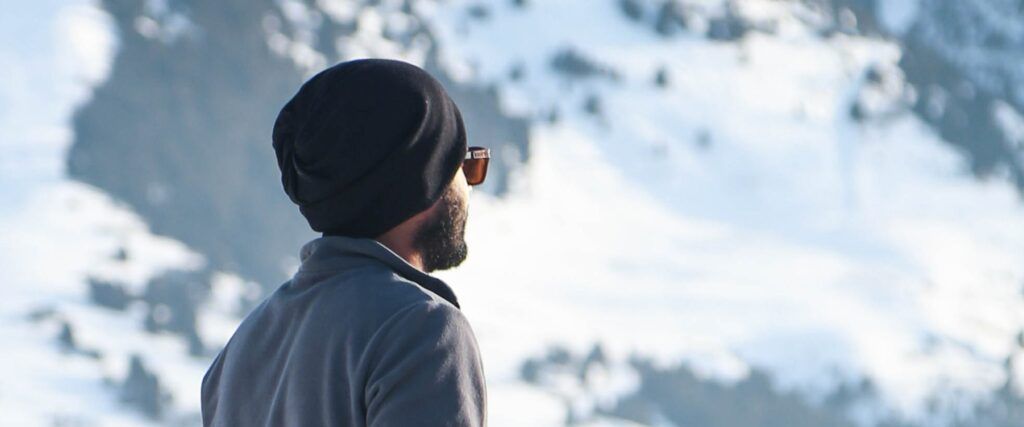 A man wearing sunglasses and a beanie looking into the snowy, alpine distance which is slightly blurred.