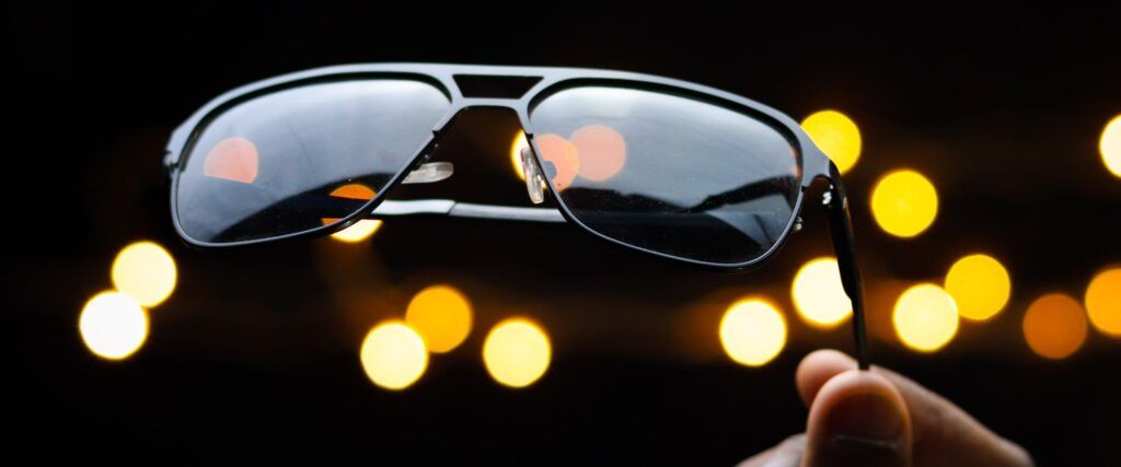 A hand holding up a pair of black sunglasses with spots of light in the background.