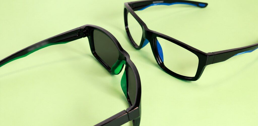 Black wraparound Harrington Sport sunglasses with tinted lenses facing black running glasses with clear lenses, both on a light-green background.
