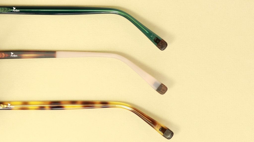 Close-up of three eyeglass temple arms in green, blush, and tortoiseshell finishes, each featuring a wooden tip, displayed against a soft yellow background.