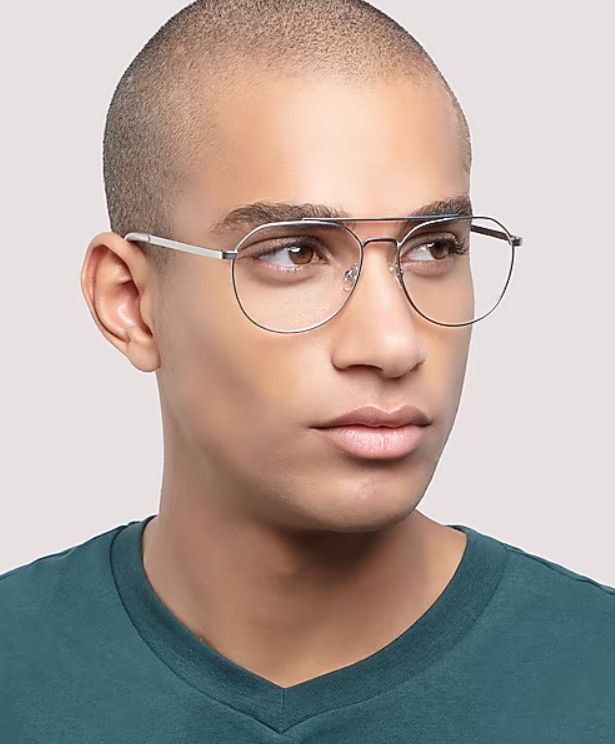 A man with a shaved head and smooth complexion wears a green top and thin silver aviator-style glasses while looking to the side. Against a soft neutral background, the minimalist portrait emphasises the lightweight metal frames and clean, modern aestheti