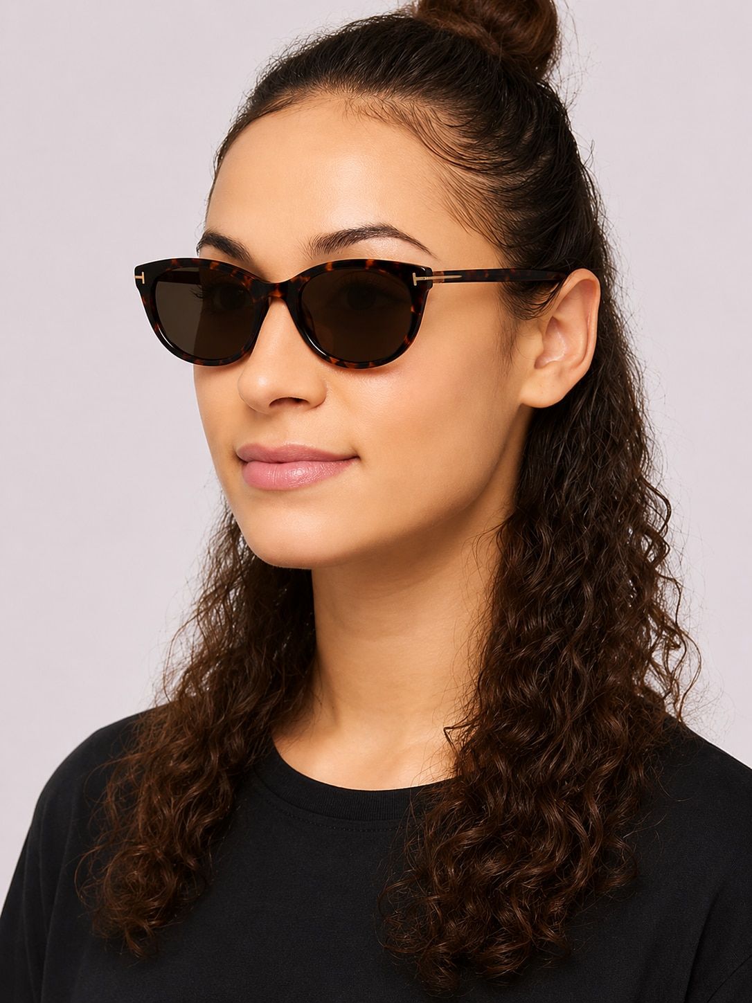 Woman with curly brown hair partly tied up, wearing a black top and oval tortoiseshell Tom Ford FT6092-B prescription sunglasses against a light background.