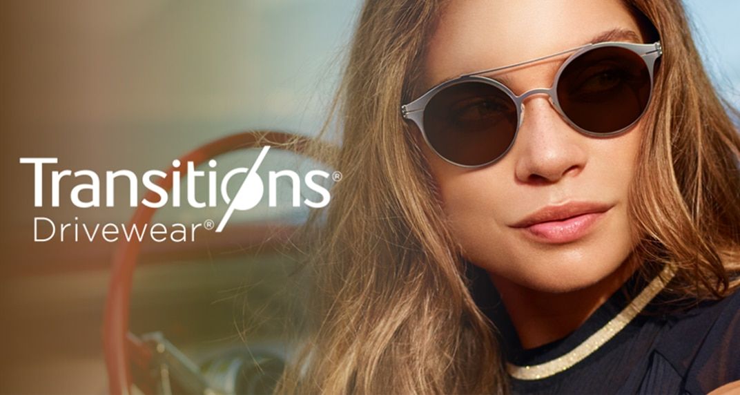 An image displaying the Transitions® Drivewear® logo in white over a background which shows a young lady behind the steering wheel of a car. She is facing away, toward the camera and has long brown hair. The lady is wearing a black top and round silver su