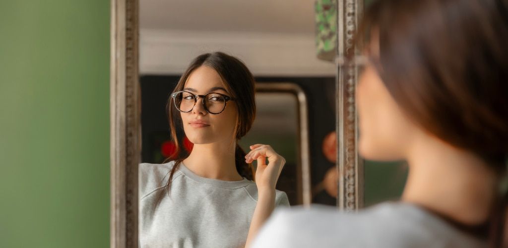 A woman with her brown hair tied loosely back wearing round tortoiseshell glasses looks at her reflection in a mirror while trying on a pair of frames at home. She's dressed casually and appears to be evaluating how the glasses look, with the mirror provi