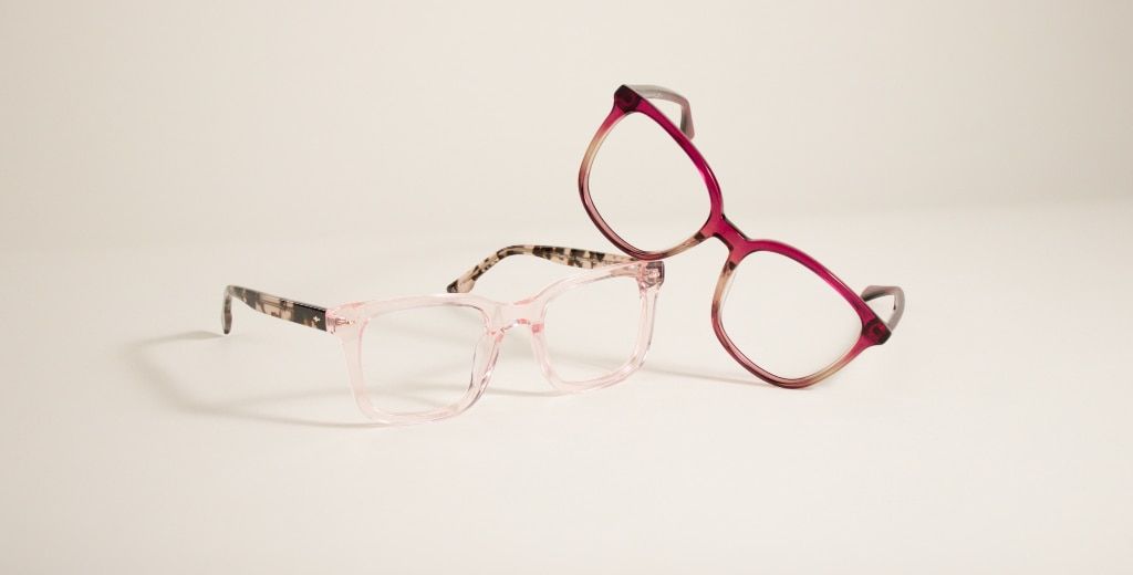Two pairs of brightly-coloured transparent frames. One has tortoiseshell temples and a clear front. The other frame is bright pink and is leaning on top of the other at an angle. The background is brightly lit and a cream colour.