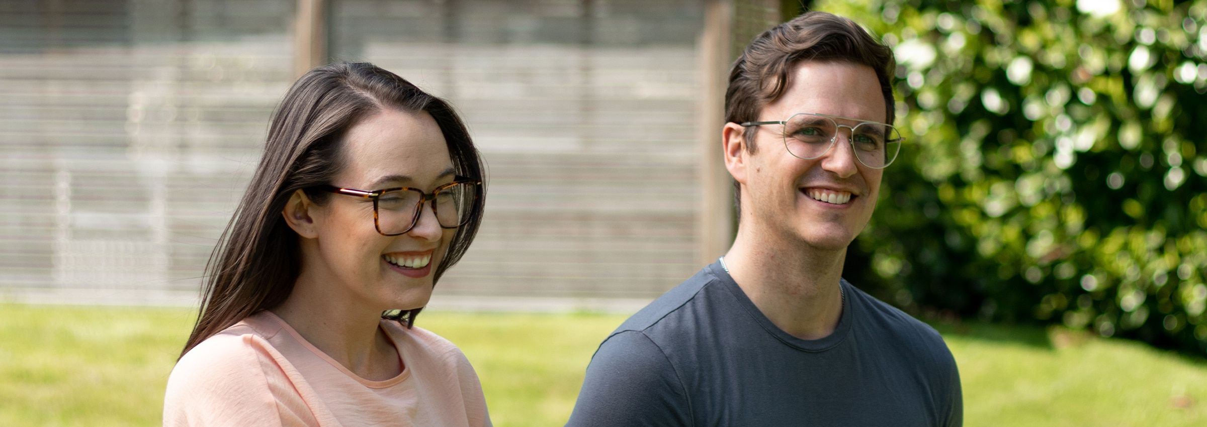 A smiling couple wearing glasses, standing outside in a garden setting as if on a date.