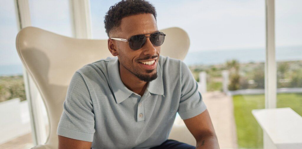 Man with short afro wearing a blue polo shirt styled with matte brown metal aviator London Retro Jenson prescription sunglasses with sleek white temples, sitting on a large chair, smiling in a bright sunlit room with an outdoor coastal view.