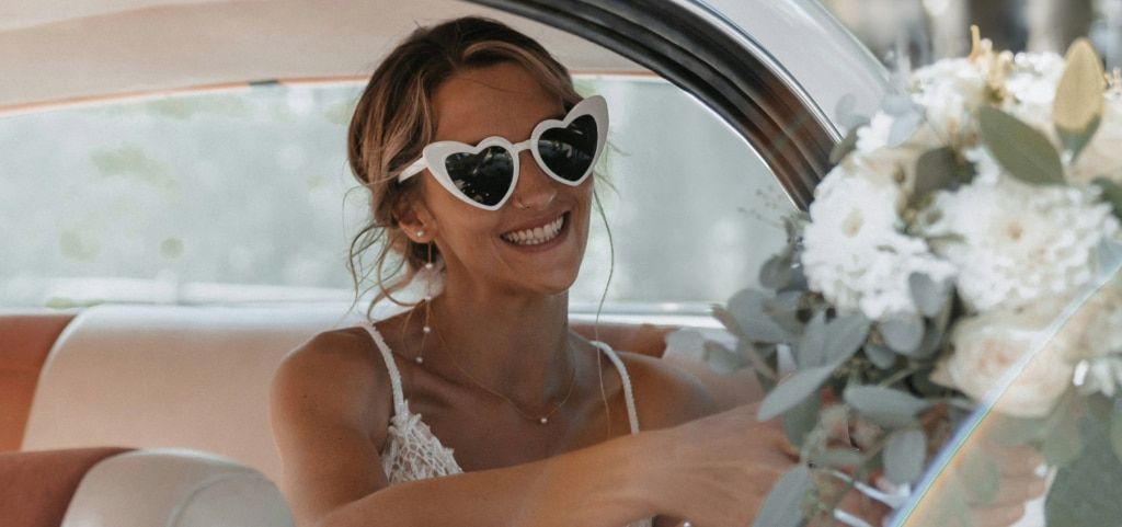 A close up of a young bride sitting in the back of a white car, handing a bouquet of white flowers to someone off camera. She has blonde wavy hair tied back with strays of hair neatly poking out. She is smiling brightly, wearing large heart-shaped sunglas