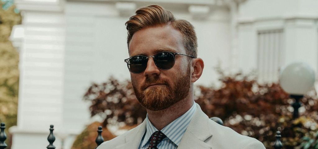 Young man with short red quiff and trimmed beard wearing round dark sunglasses, a striped shirt, red polka-dot tie and cream blazer, standing outdoors near a black iron gate leading to a private garden behind him.
