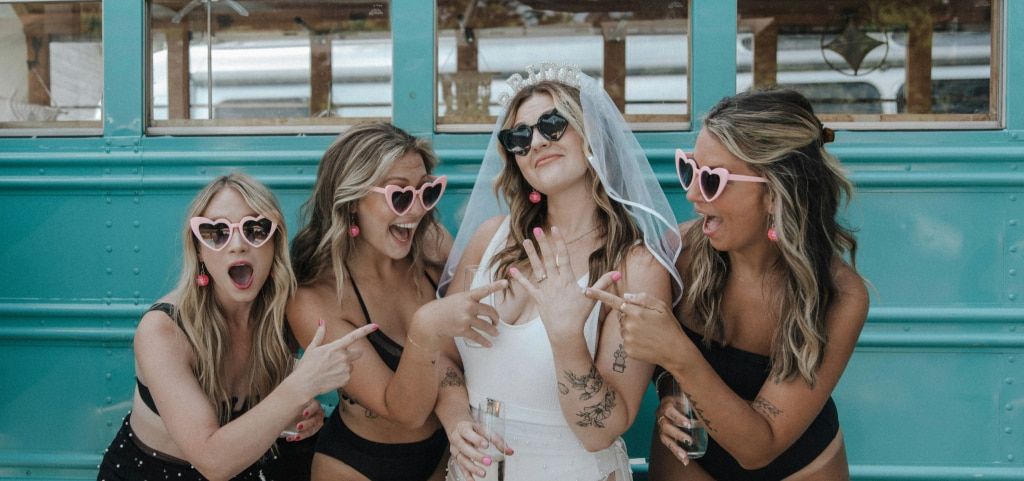 Bride-to-be in a veil and white swimsuit wearing black heart-shaped sunglasses, showing her engagement ring as friends in matching pink heart sunglasses point excitedly beside a blue bus.
