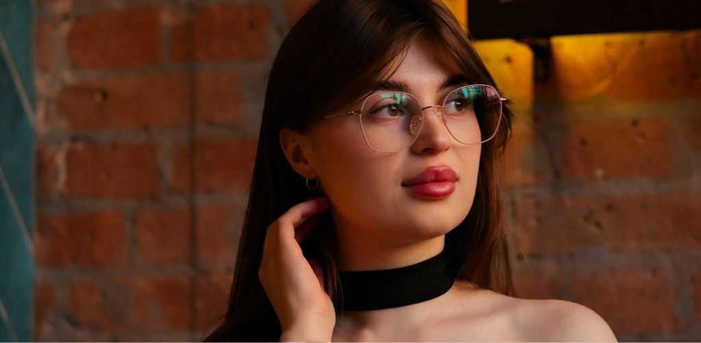 A woman with long straight brown hair wearing large round metal eyeglasses, pink lipstick, and a black top with a choker neck looks thoughtfully to the side, her hand resting near her neck, with warm light and an exposed brick wall in the background.