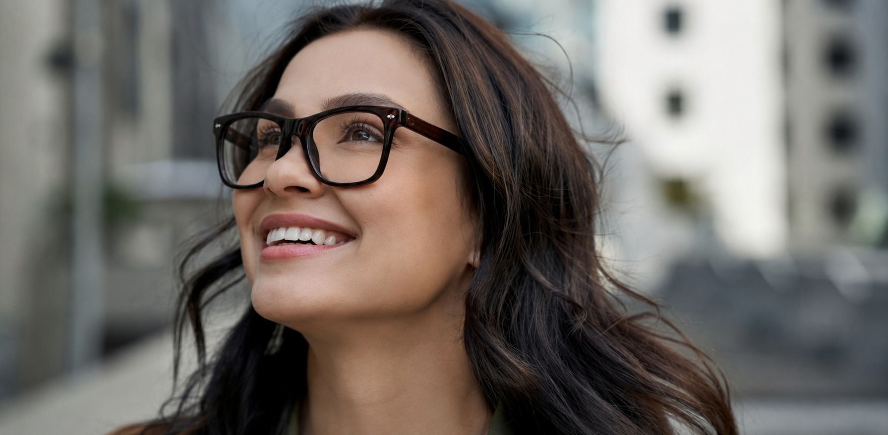 Close-up of a smiling woman with wavy brown hair wearing dark-brown wayfarer-style glasses with metal pin detailing in an urban setting. The clear, scratch-free lenses highlight the durability of scratch-resistant eyewear and support clear vision for ever
