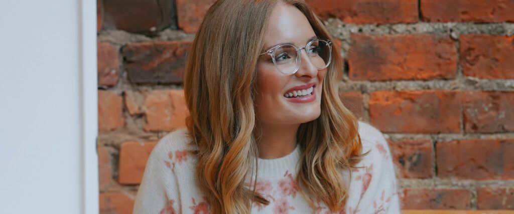 Close-up of a woman with long, wavy blonde hair smiling brightly and looking off to the side. She wears transparent round acetate glasses and a white floral jumper, with a brick wall behind her.