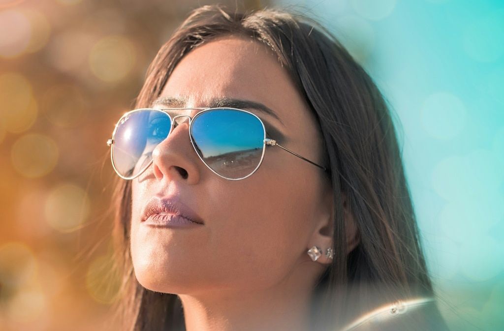 Close-up of a woman with straight brown hair wearing Ray-Ban aviator sunglasses in bright sunlight
