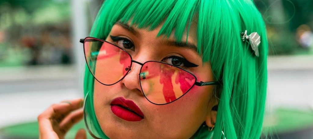 A young woman with green hair wearing thin black cat eye glasses with almost triangular lenses with a pink tint