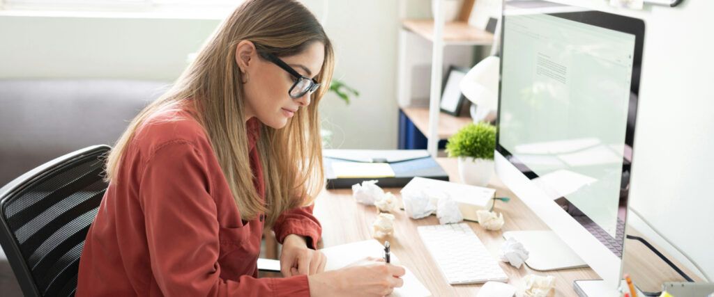 A young lady with long straight blonde hair wearing a red shirt and a pair of black square acetate glasses. The lady is sitting at a work desk writing on a piece of paper. There is an iMac computer screen and keyboard in front of her, as well as various s