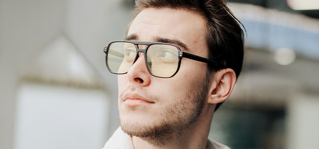 A close up of a young man in a bright blurred-out setting. He has short brown hair and is wearing a pair of black pilot-style glasses. The man is looking up to his right.