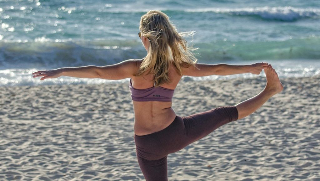 A blonde woman in light gym clothes doing yoga on a sandy beach