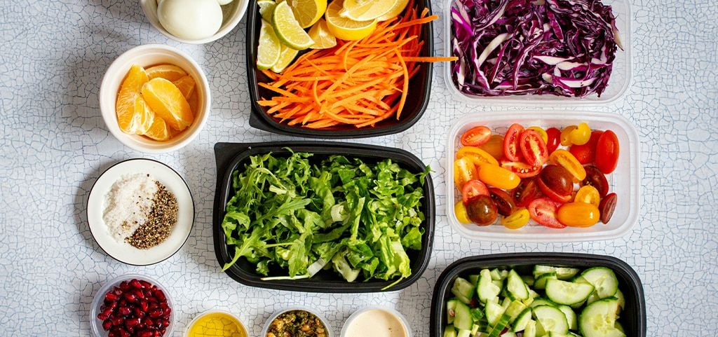 A selection of healthy foods laid out in bowls and containers, including boiled eggs, chopped carrots and limes, shredded red cabbage, chopped tomatoes, chopped cucumber, shredded lettuce, tangerine pieces, pomegranate seeds and several spices and sauces.