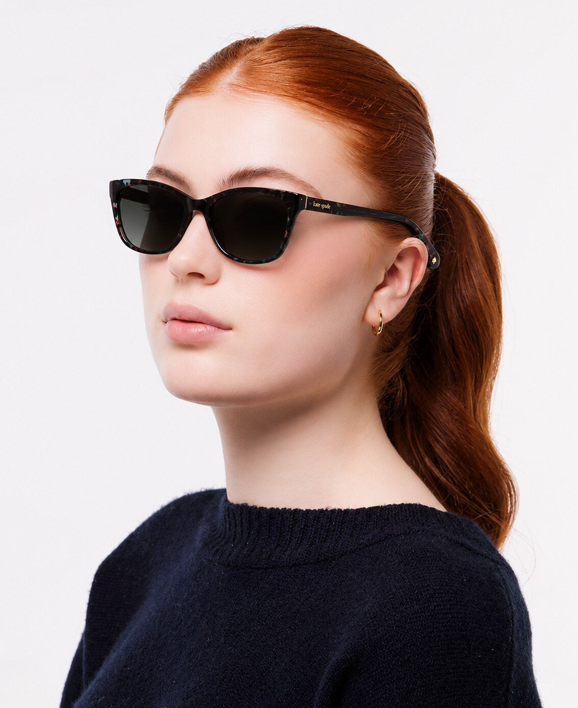 A woman with red hair tied neatly back in a ponytail wears small rectangular sunglasses with a dark blue jumper, standing at angle against a plain background.