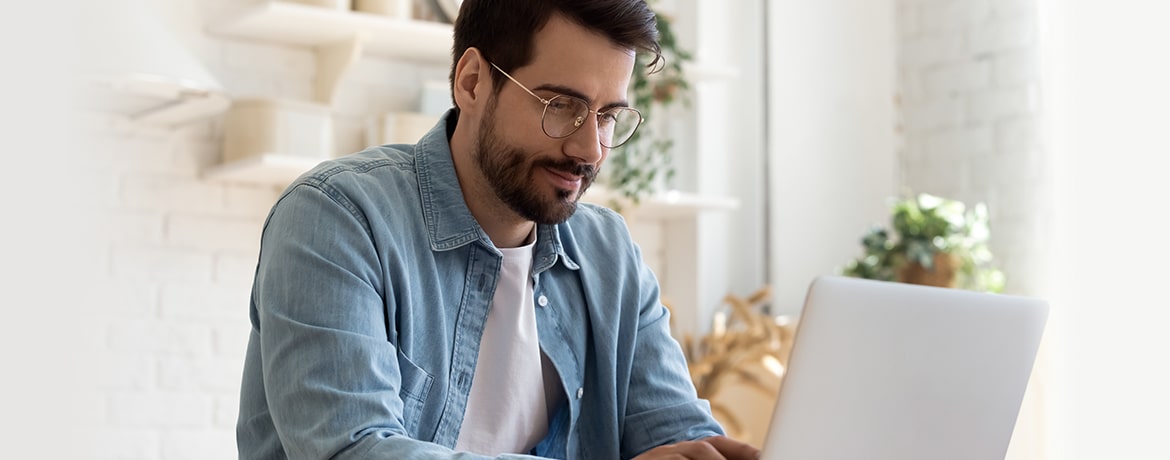 A young man with a beard and wearing glasses browses for new frames on his laptop from the comfort of his home.