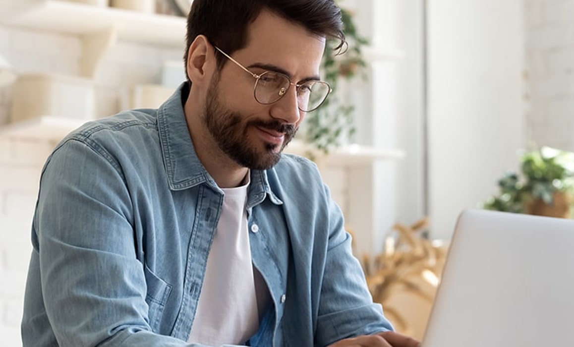 A young man with a beard and wearing glasses browses for new frames on his laptop from the comfort of his home.