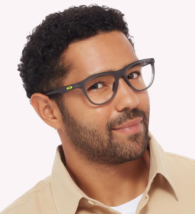 Image shows a man wearing grey sport glasses frames with a light pink background.