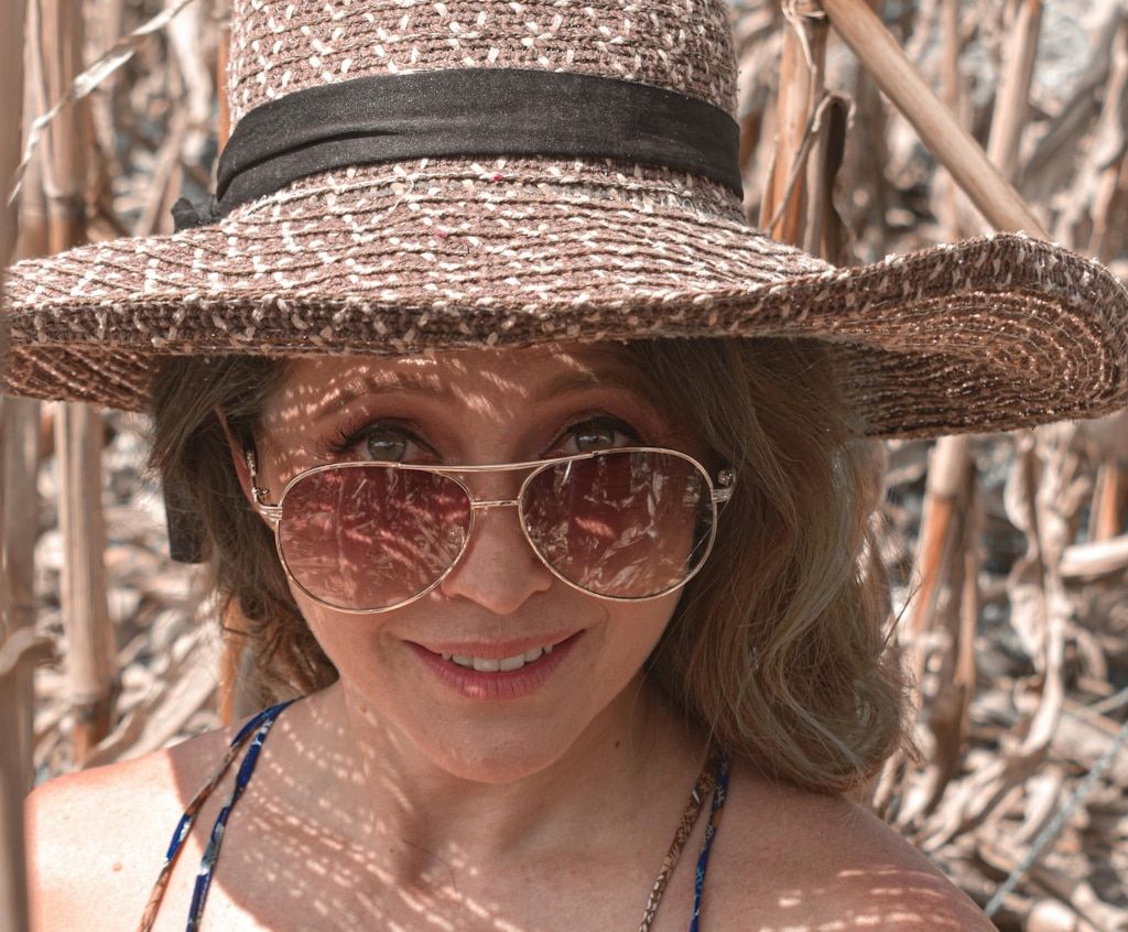 A blonde woman with a straw hat peeking over the rims of her aviator sunglasses with brown gradient lenses