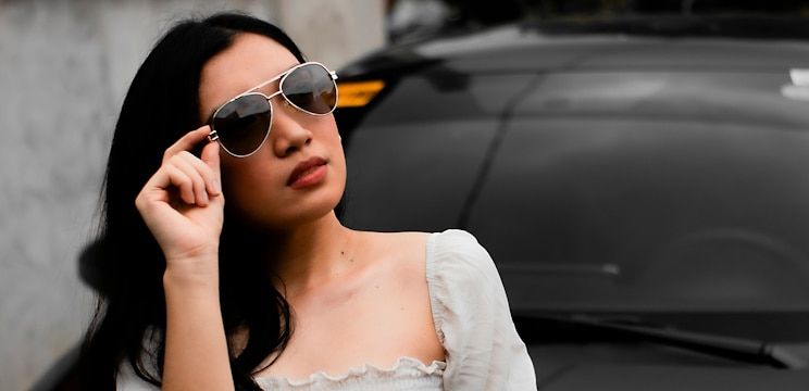 A woman with long black hair putting on aviator sunglasses in front of her car