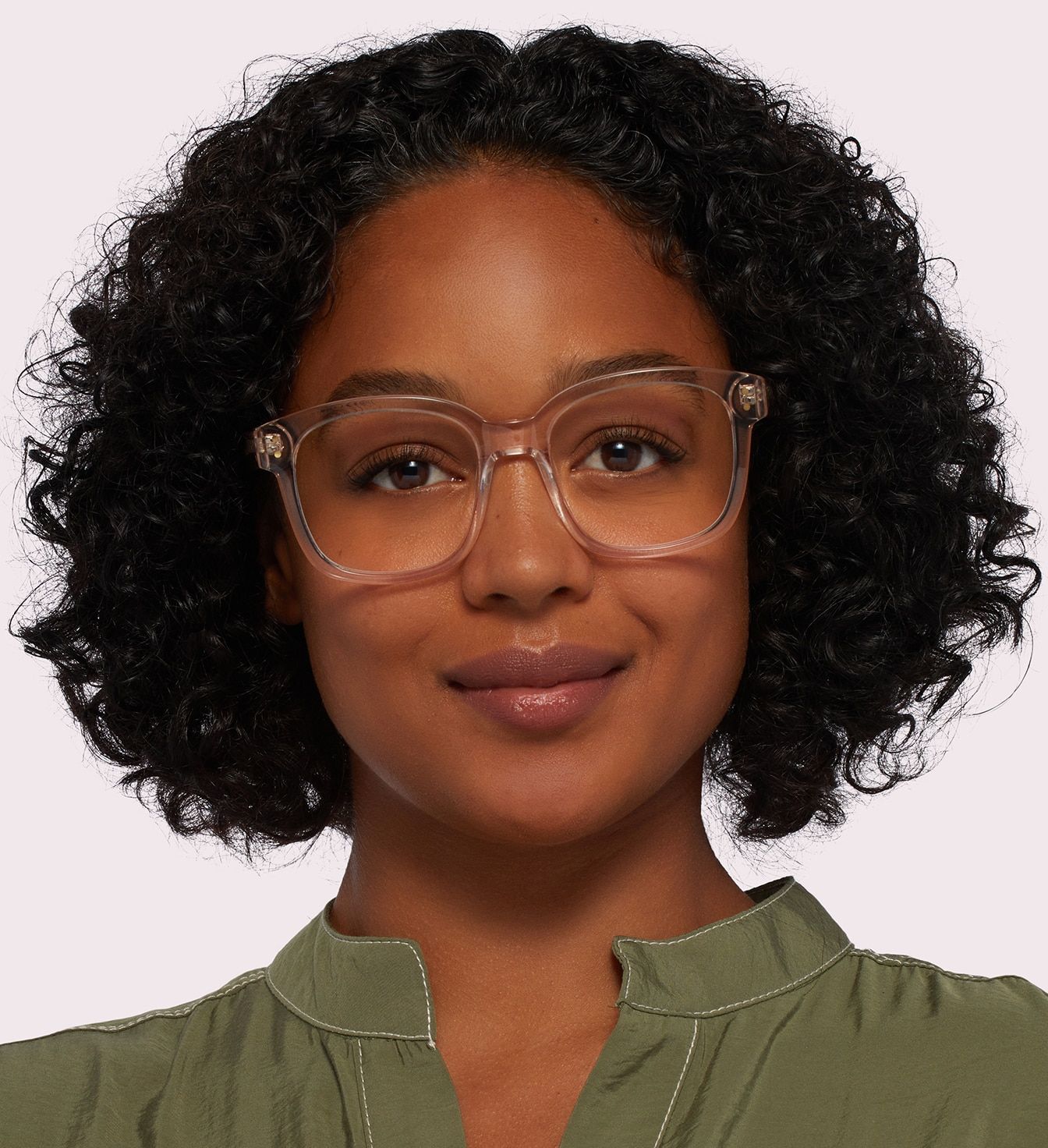 A close-up of a young lady with thick curly black hair. She is wearing a pair of large transparent square glasses and a green collarless blouse.