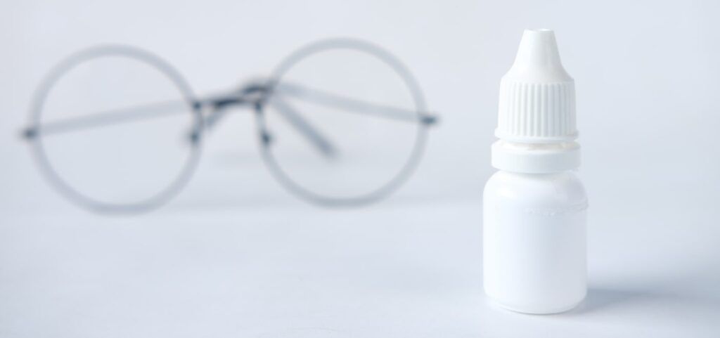 A close-up of a white eye drop bottle, with a pair of round metal glasses softly blurred in the white background.